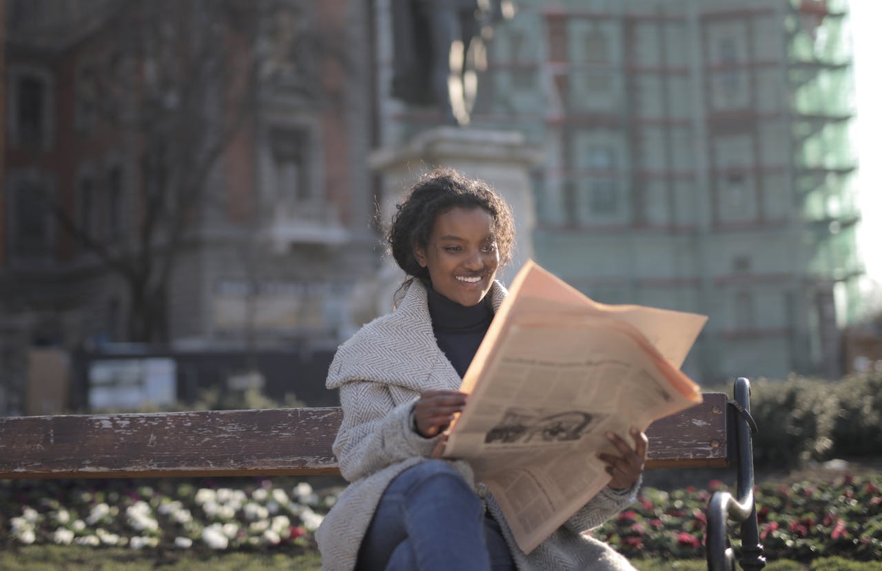 Cheerful woman enjoys reading a newspaper while seated on a park bench during a sunny day.